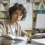 Étudiant(e) concentré(e) avec des lunettes, levant les yeux de ses livres dans une bibliothèque ou une salle d'étude remplie de formules.