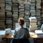 Une personne travaille devant une grande pile de livres, concentrée sur son ordinateur, entourée de documents