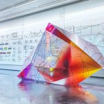 Grande structure géométrique 3D (polyèdre) aux reflets colorés, exposée dans un laboratoire de design ou d'architecture moderne avec des schémas en arrière-plan.